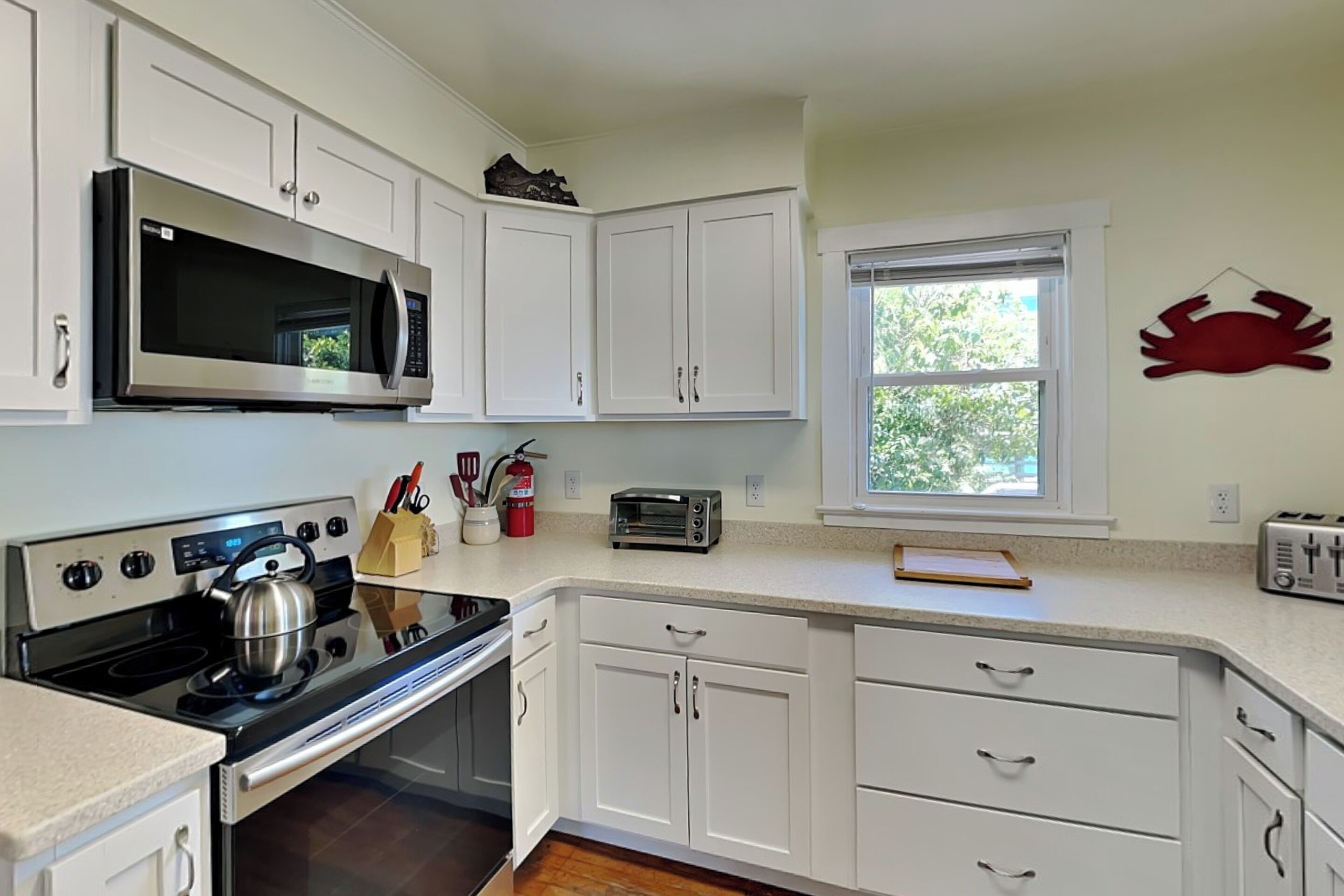 A kitchen with white cabinets, a microwave, a stove, a toaster, and a window with a view of greenery.