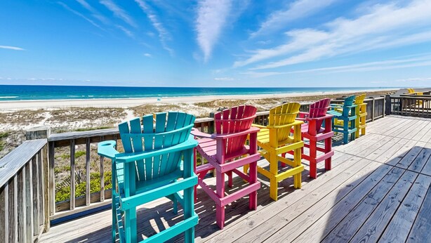Une terrasse en bois avec des fauteuils Adirondack aux couleurs vives, donnant sur une plage.