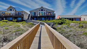 Une promenade en bois mène à une propriété située en bord de mer, avec des maisons à plusieurs étages, un ciel dégagé et un paysage sablonneux.