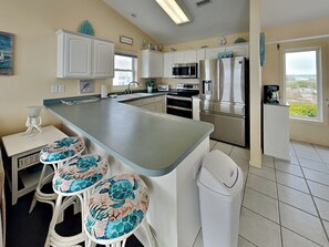 A kitchen with white cabinets, a large island with stools, stainless steel appliances, and a view through the window.