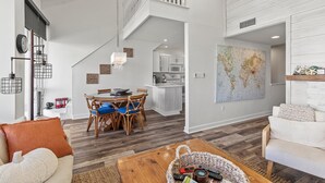 Un salón amplio con una mesa de comedor, sillas, una mesa de centro y un sofá. Se ve una cocina al fondo, con armarios y un microondas.
