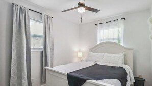 Een slaapkamer met een wit bed, een plafondventilator, een raam met jaloezieën en een nachtkastje met een lamp.
