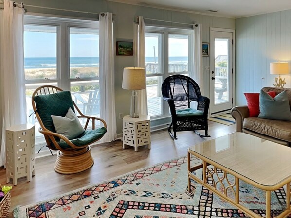 A living room with a view of the beach, wicker chairs, a glass coffee table, and a patterned rug.