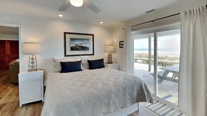 A bedroom with a large bed, two bedside tables, a ceiling fan, a framed picture, and a view of the beach through a sliding glass door.