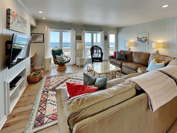 A living room with a TV, two sofas, a coffee table, and a view of the ocean.