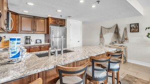 A kitchen with granite countertops, wooden cabinets, a stainless steel refrigerator, and a bar area with bar stools. - Sea Club #104 - Casa con Vista (Indian Shores)