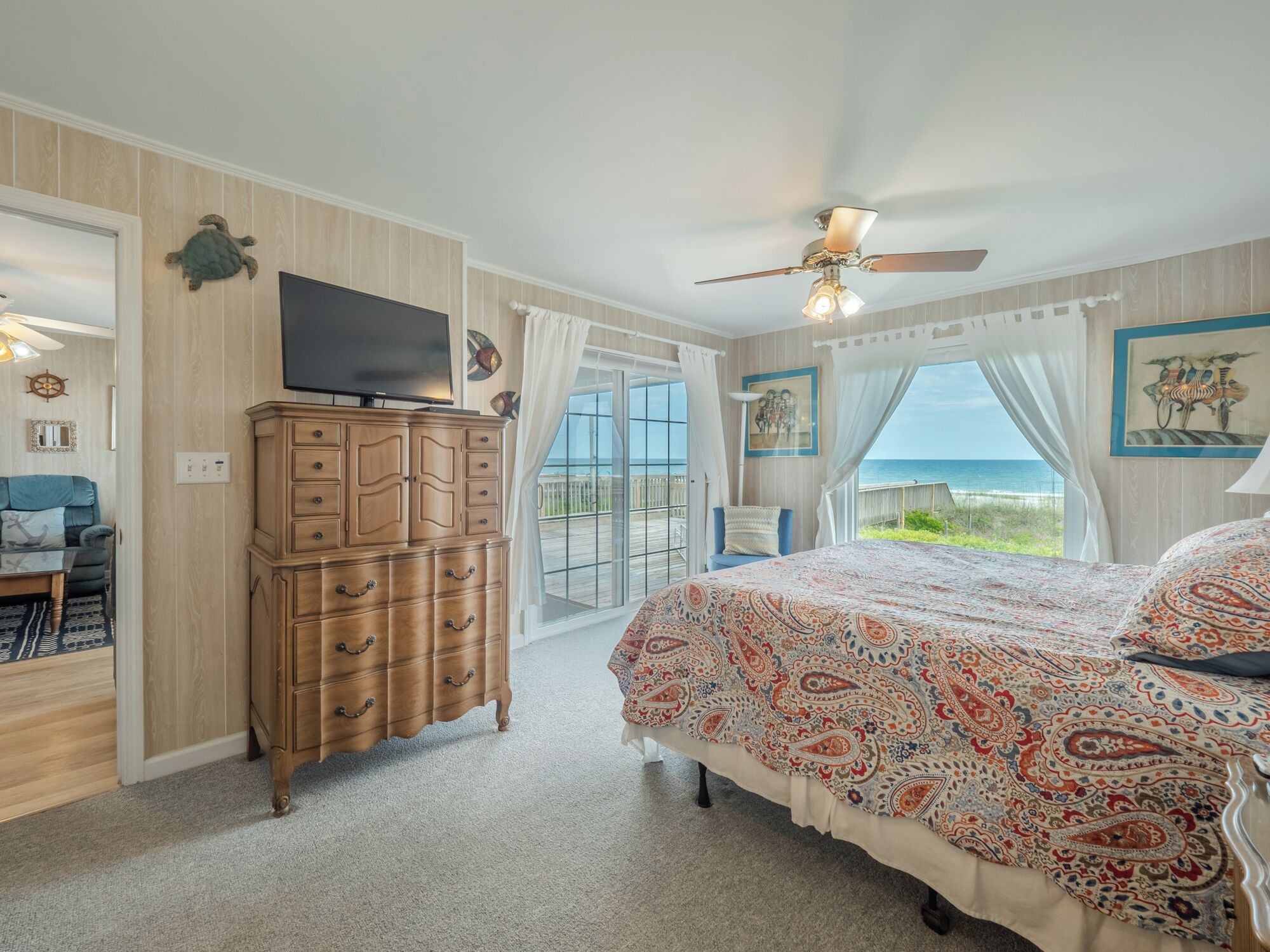 A bedroom with a large bed, a wooden dresser, a television, and a view of the ocean through a glass door.