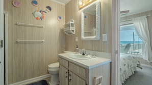 A bathroom with a wooden wall, a white sink, a mirror with lights, and a view of the beach through the window.