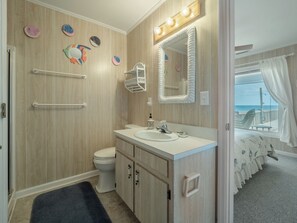A bathroom with a wooden wall, a white sink, a mirror with lights, and a view of the beach through the window.