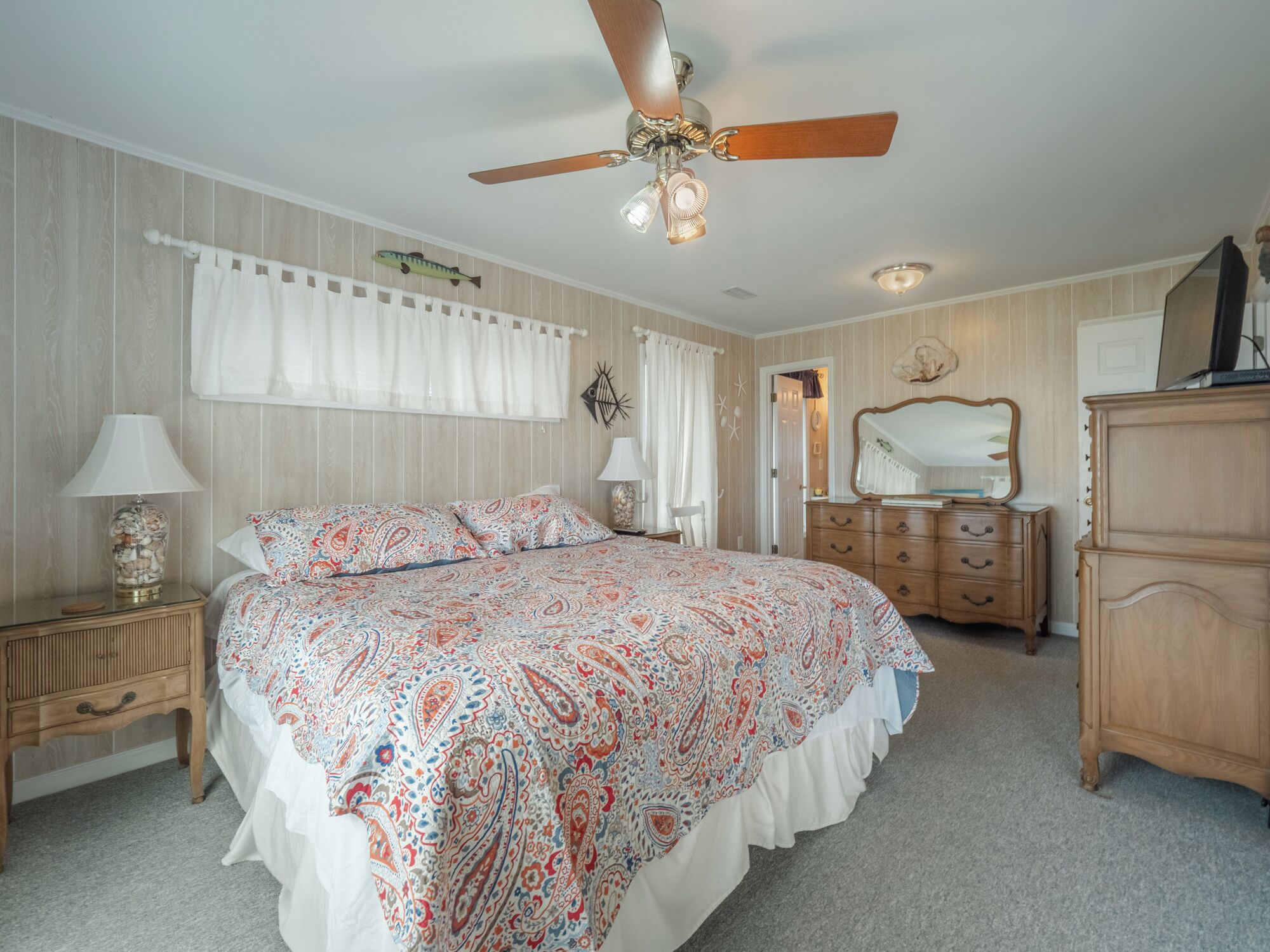 Ein Schlafzimmer mit einem Bett, einer Kommode, einem Nachttisch, einem Deckenventilator und einem Fenster mit Vorhängen.