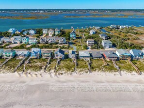An aerial view of a coastal residential area with houses connected by walkways, a beach in the foreground, and a large body of water in the background. - Blue Crab by Topsail Realty (Holly Ridge)