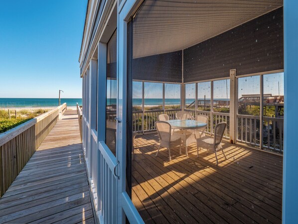 A wooden deck with a view of the ocean, a covered outdoor seating area with chairs and a table, and a large window overlooking the beach.