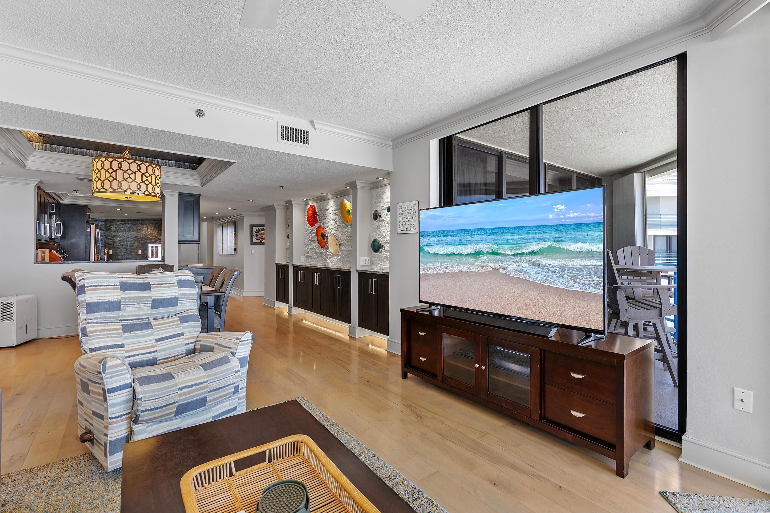 Ein geräumiges Wohnzimmer mit einem großen Flachbildfernseher, der eine Strandansicht zeigt, einem bequemen Sofa und einer Holzkommode.