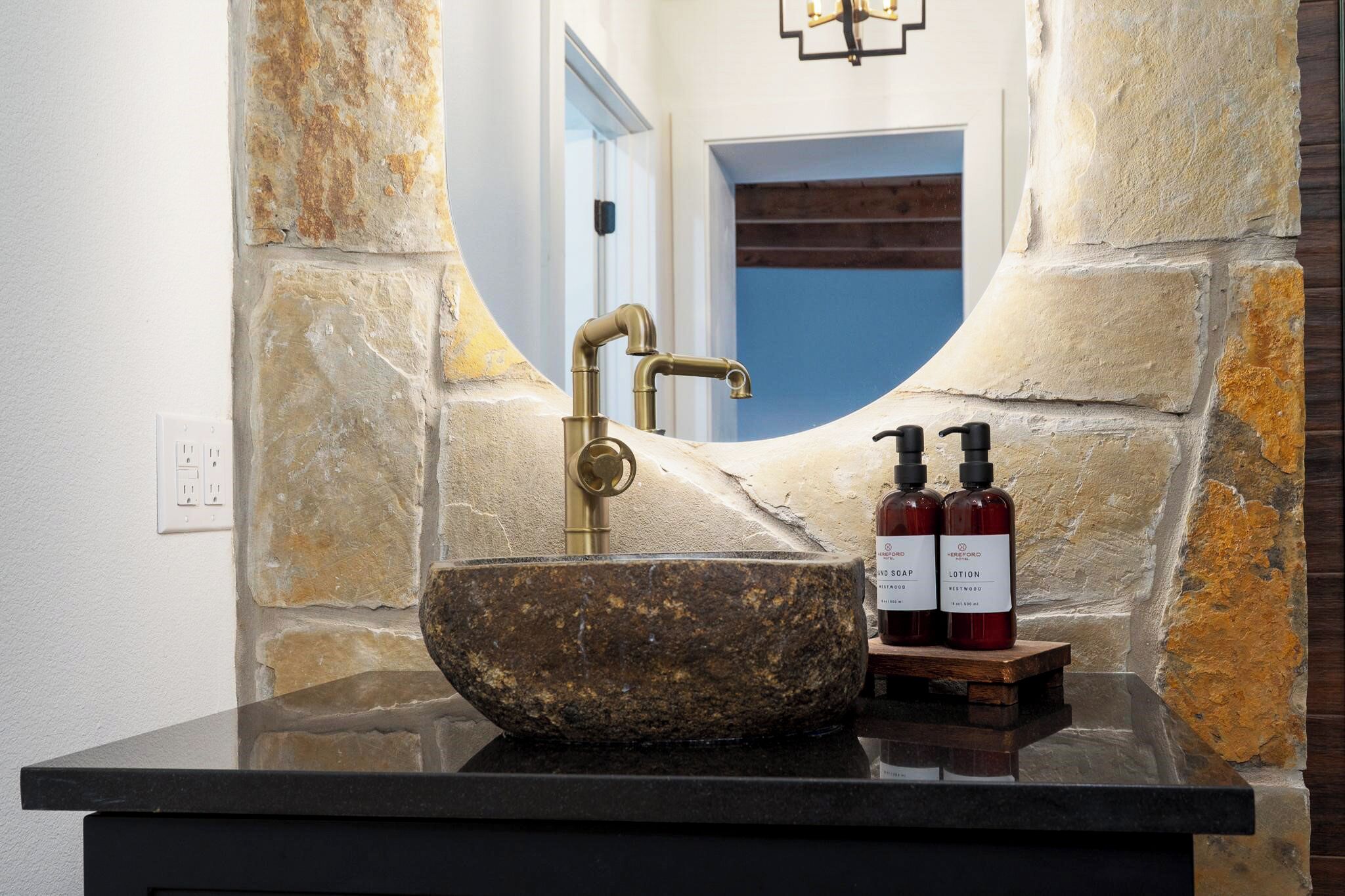 A stone sink with a brass faucet, two bottles of lotion, and a mirror in a stone-walled bathroom.