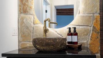 A stone sink with a brass faucet, two bottles of lotion, and a mirror in a stone-walled bathroom.