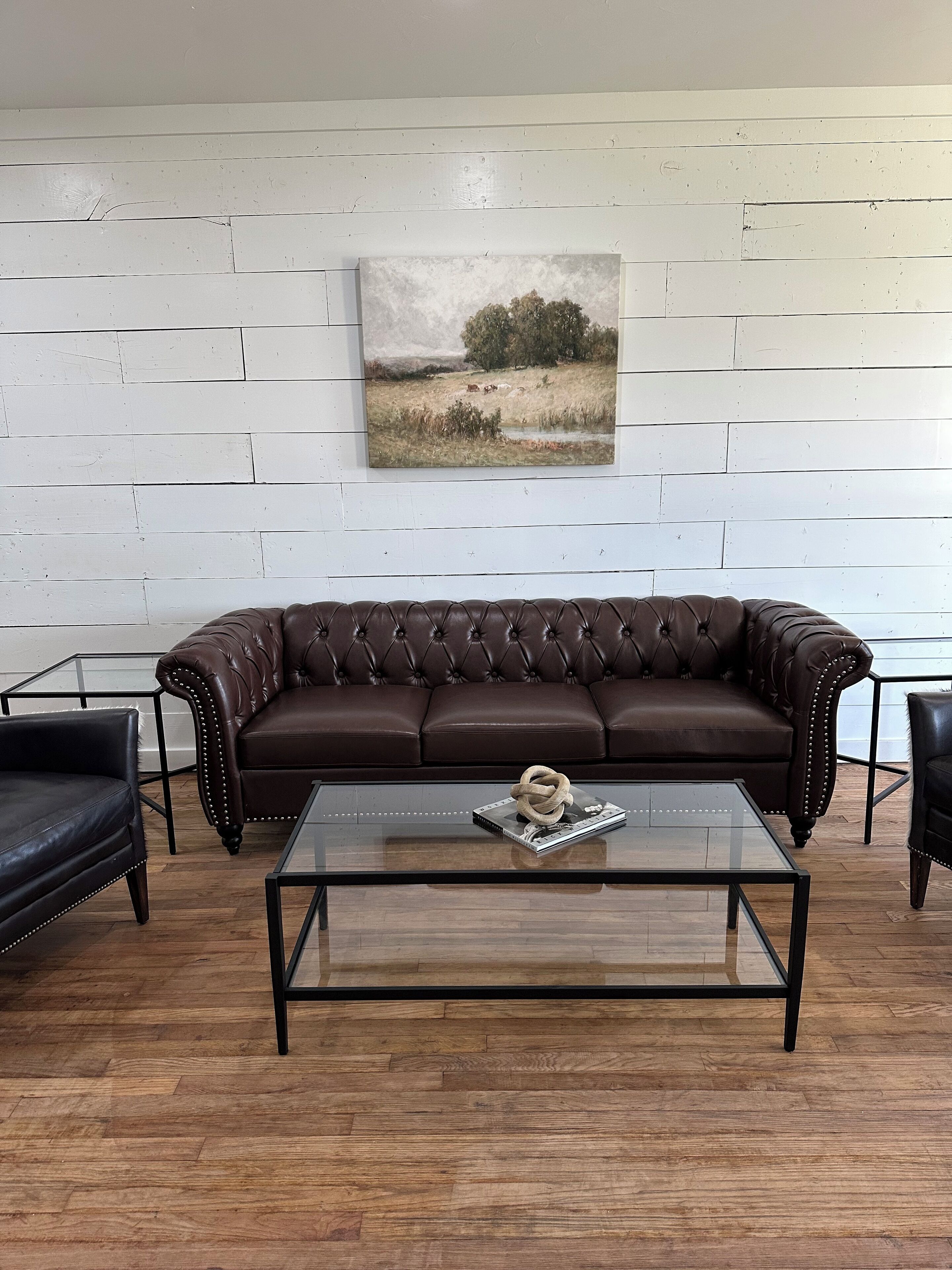 Een bruine leren bank tegen een witte houten wand, met daarboven een landschap. Een glazen salontafel met een tijdschrift en een touwsculptuur bevindt zich ervoor.