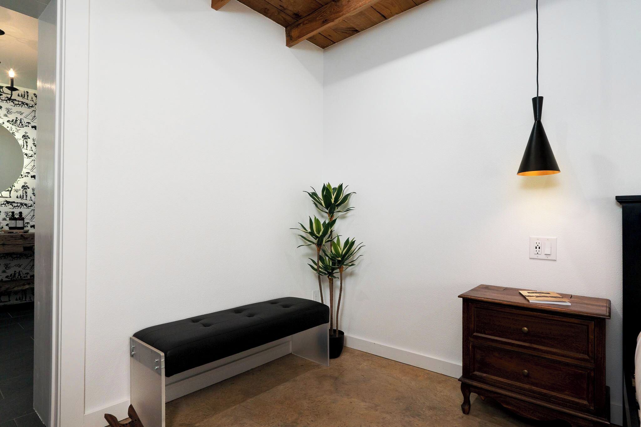 A corner room with a bench, a potted plant, a wooden dresser, and a hanging light fixture.