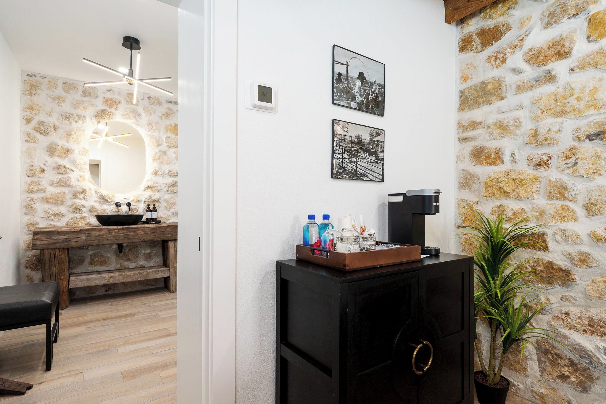 A room with a stone wall, a wooden bench, a black cabinet with a plant, and a mirror with a light fixture.