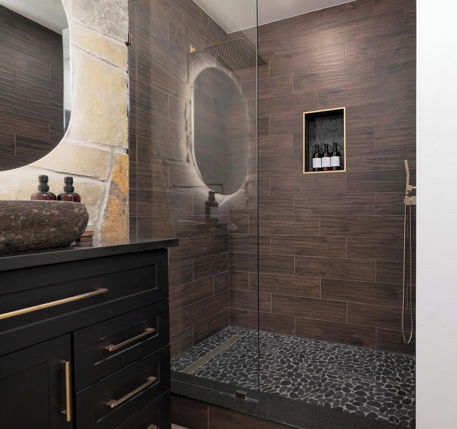 A modern bathroom with a stone sink, dark wood tiles, a glass shower enclosure, and a small shelf with bottles.