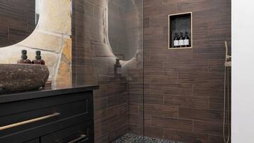 A modern bathroom with a stone sink, dark wood tiles, a glass shower enclosure, and a small shelf with bottles.