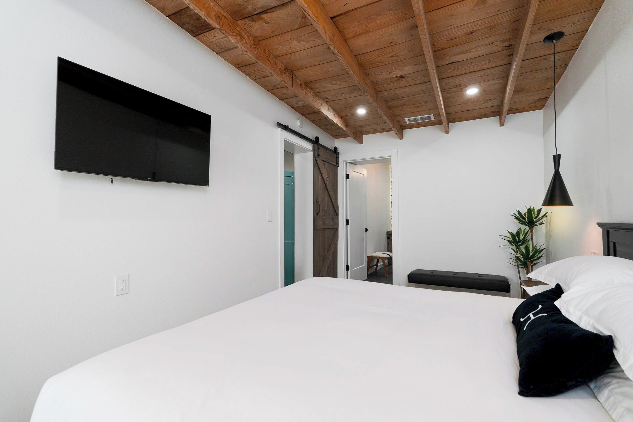 Een slaapkamer met een groot bed, een aan de muur bevestigde televisie, een schuifdeur, een bankje en een hanglamp.