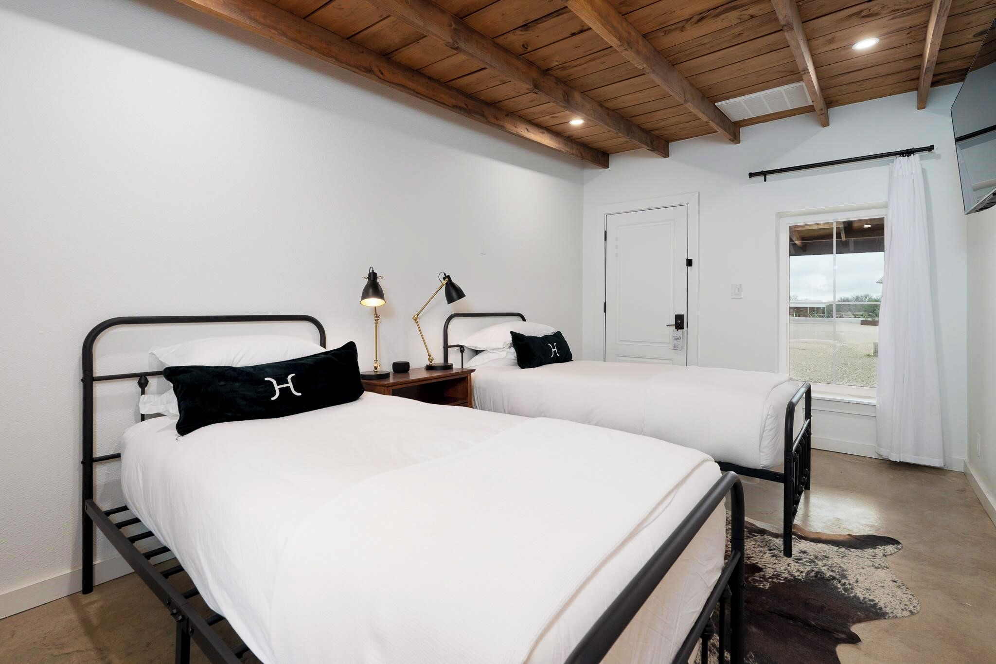A bedroom with two beds, a wooden ceiling, a window with curtains, and a door.