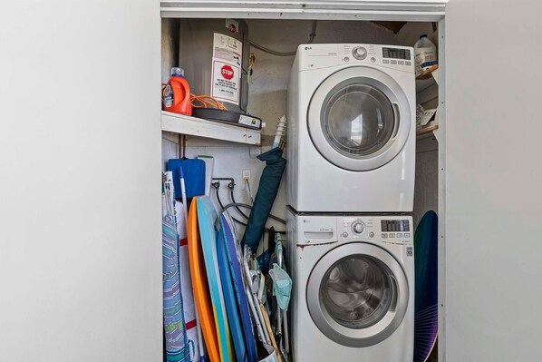 A laundry room with two stacked washing machines, a shelf with cleaning supplies, and various cleaning tools hanging on the wall.
