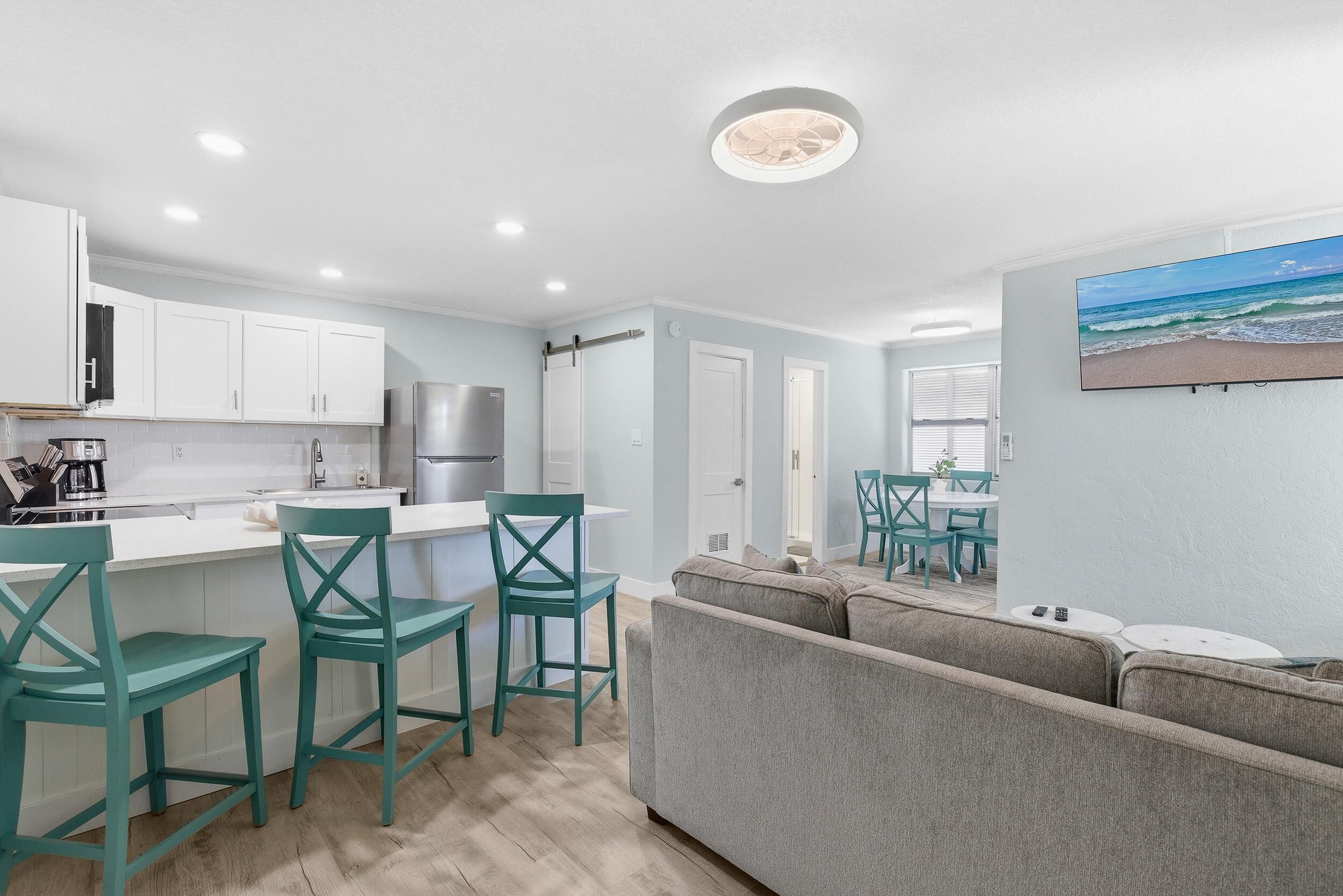 A modern kitchen with white cabinets, teal chairs, and a grey sofa in a bright room with a ceiling fan and a beach painting.