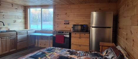 A compact wooden cabin with a kitchenette, a bed with a patterned blanket, and a view of the outdoors through a window.