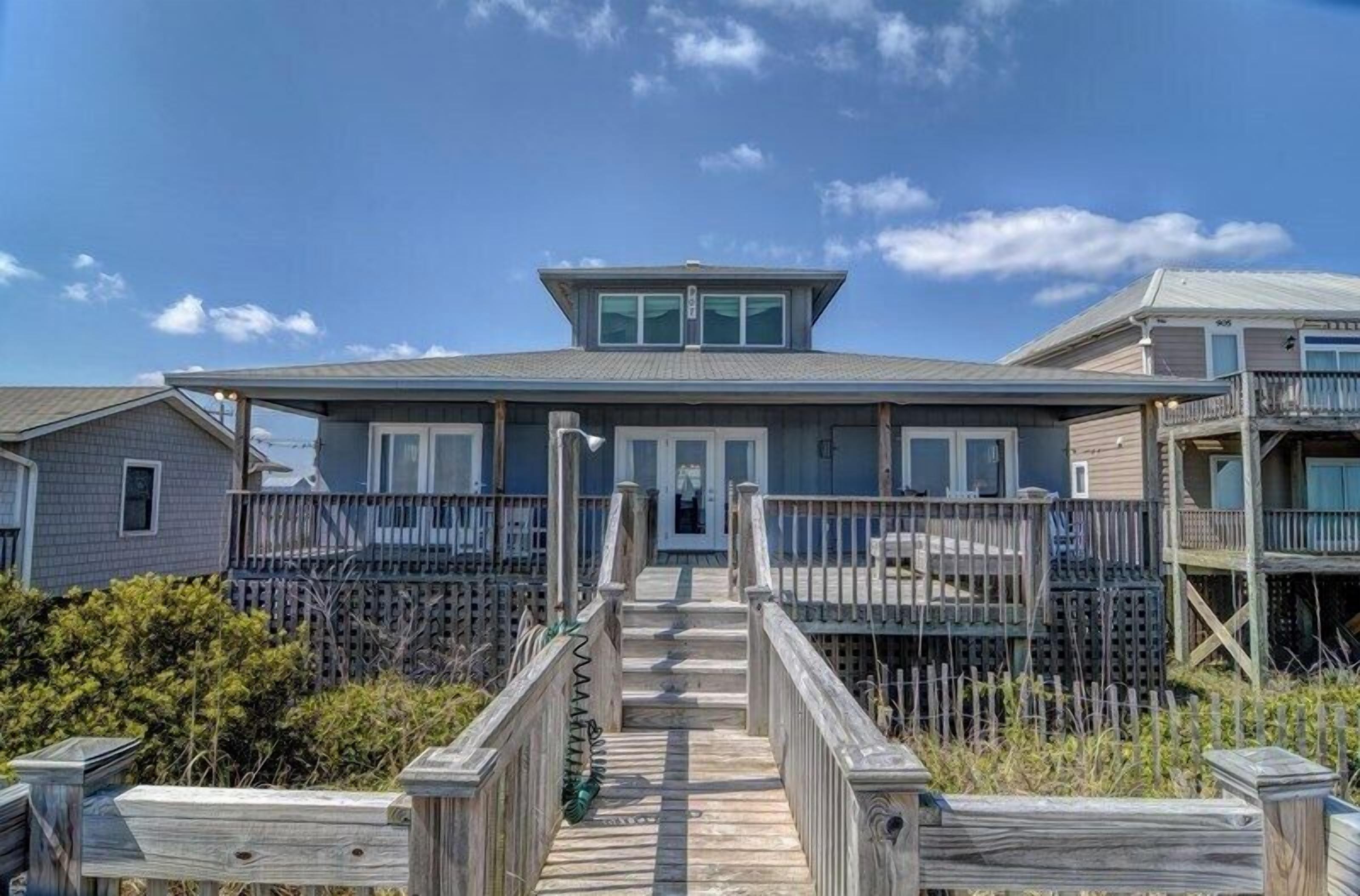 A beachfront property with a wooden deck leading to a house with a covered porch and a second-story balcony.