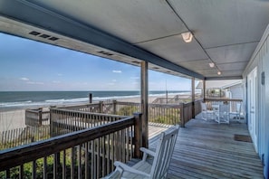 A covered deck with a view of the ocean, wooden railings, and white chairs.
