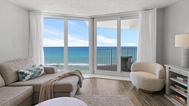 Una sala de estar moderna con un ventanal grande con vista al océano, un sofá beige, un sillón blanco y una estantería.