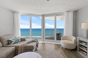 A modern living room with a large window overlooking the ocean, a beige sofa, a white armchair, and a bookshelf. - The Shores Club 1105 (Daytona Beach Shores)