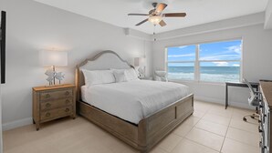 A bedroom with a bed, a nightstand, a desk, and a window with a view of the ocean. - Aruba 206 (Daytona Beach Shores)