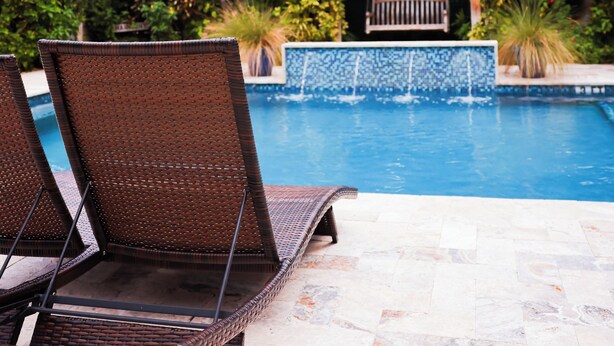 Un área junto a la piscina con sillones de mimbre, una piscina con baldosas y plantas tropicales.