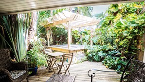 Una terraza de madera con pérgola, zona de estar y abundante vegetación.
