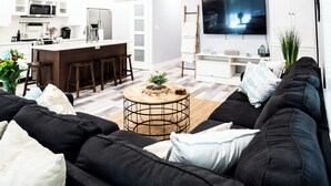 Una sala moderna con un sofá negro, una mesa redonda de centro y un televisor. Hay una zona de cocina con armarios blancos y un microondas.