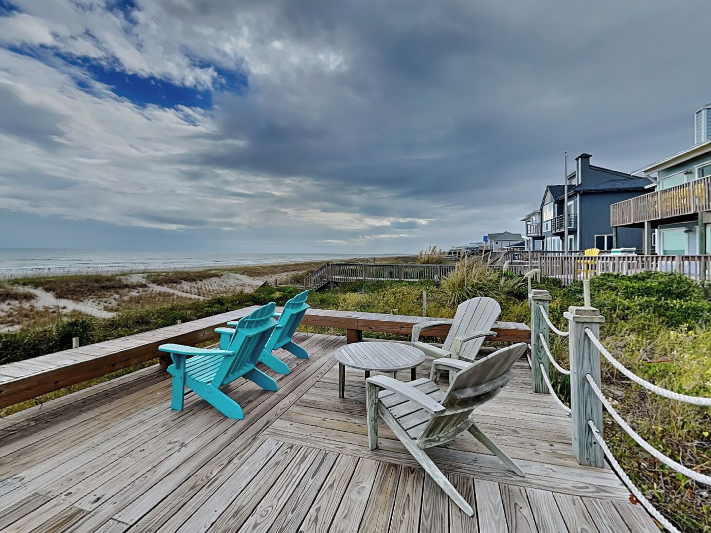 A wooden deck with chairs overlooking a beach and a house in the background.