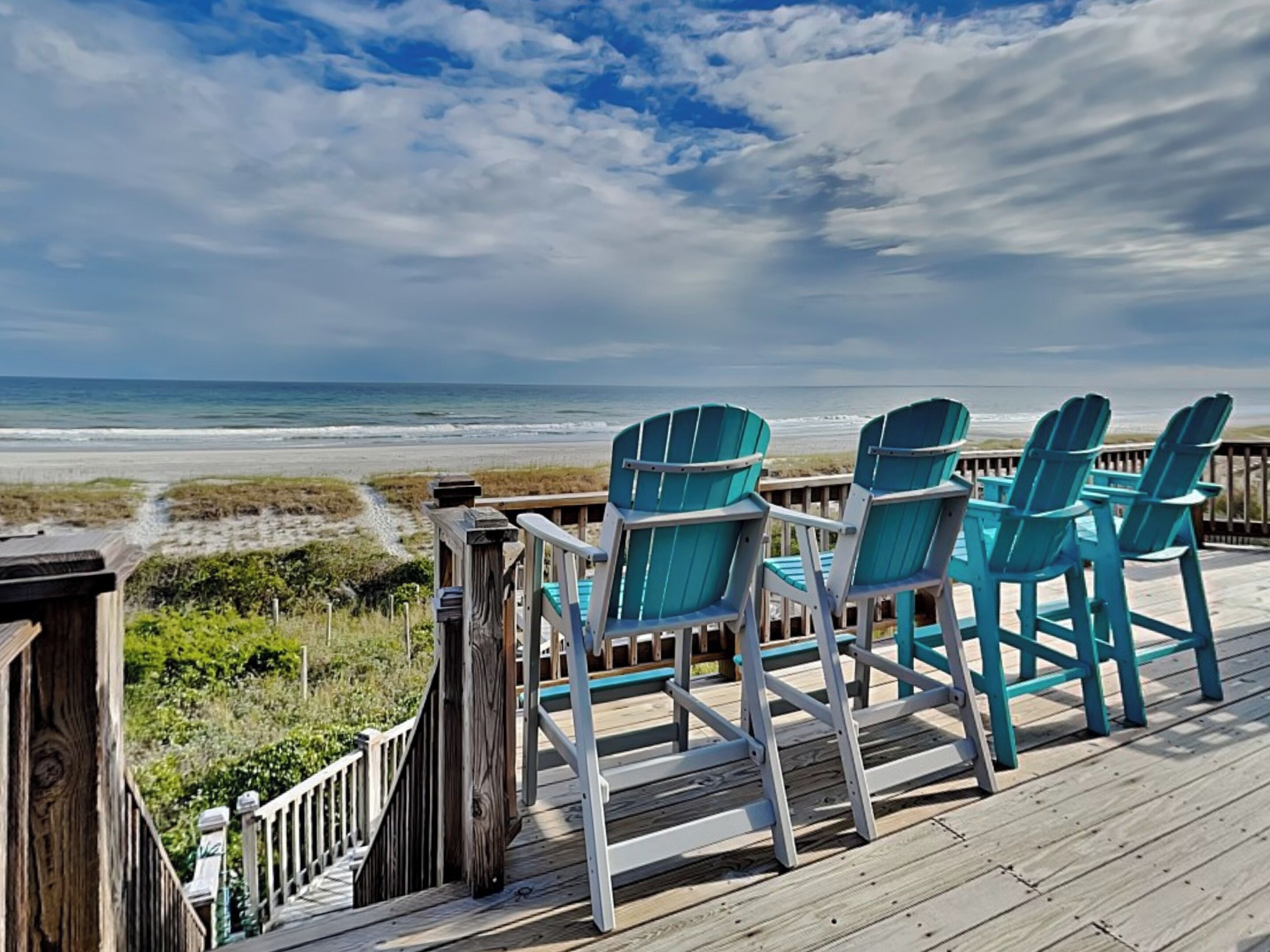 Une terrasse en bois avec des fauteuils Adirondack turquoise donnant sur une plage.