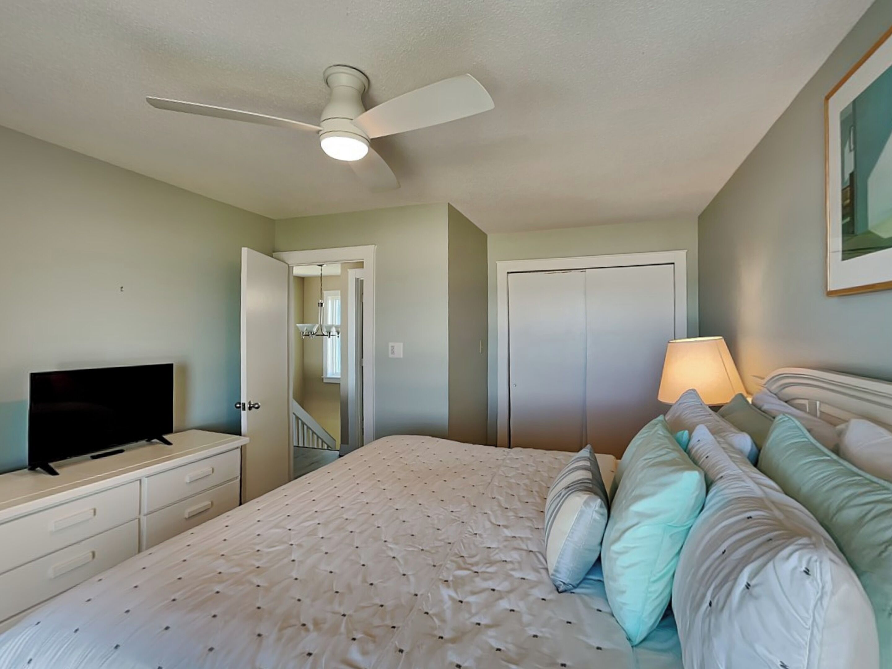 A bedroom with a bed, a television on a dresser, a ceiling fan, and a door leading to a hallway.