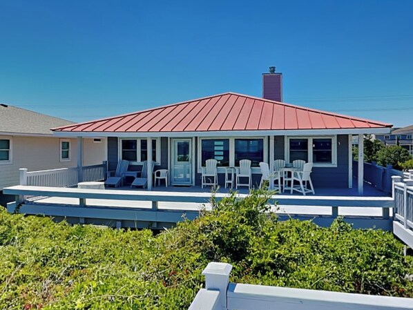 A two-story house with a red roof, a covered porch with white chairs and tables, and a small garden area in front. - A Beach Pearle by Topsail Realty (Holly Ridge)