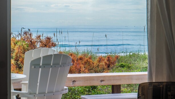 Une vue depuis une fenêtre donnant sur l’océan, avec une terrasse en bois et une chaise blanche.