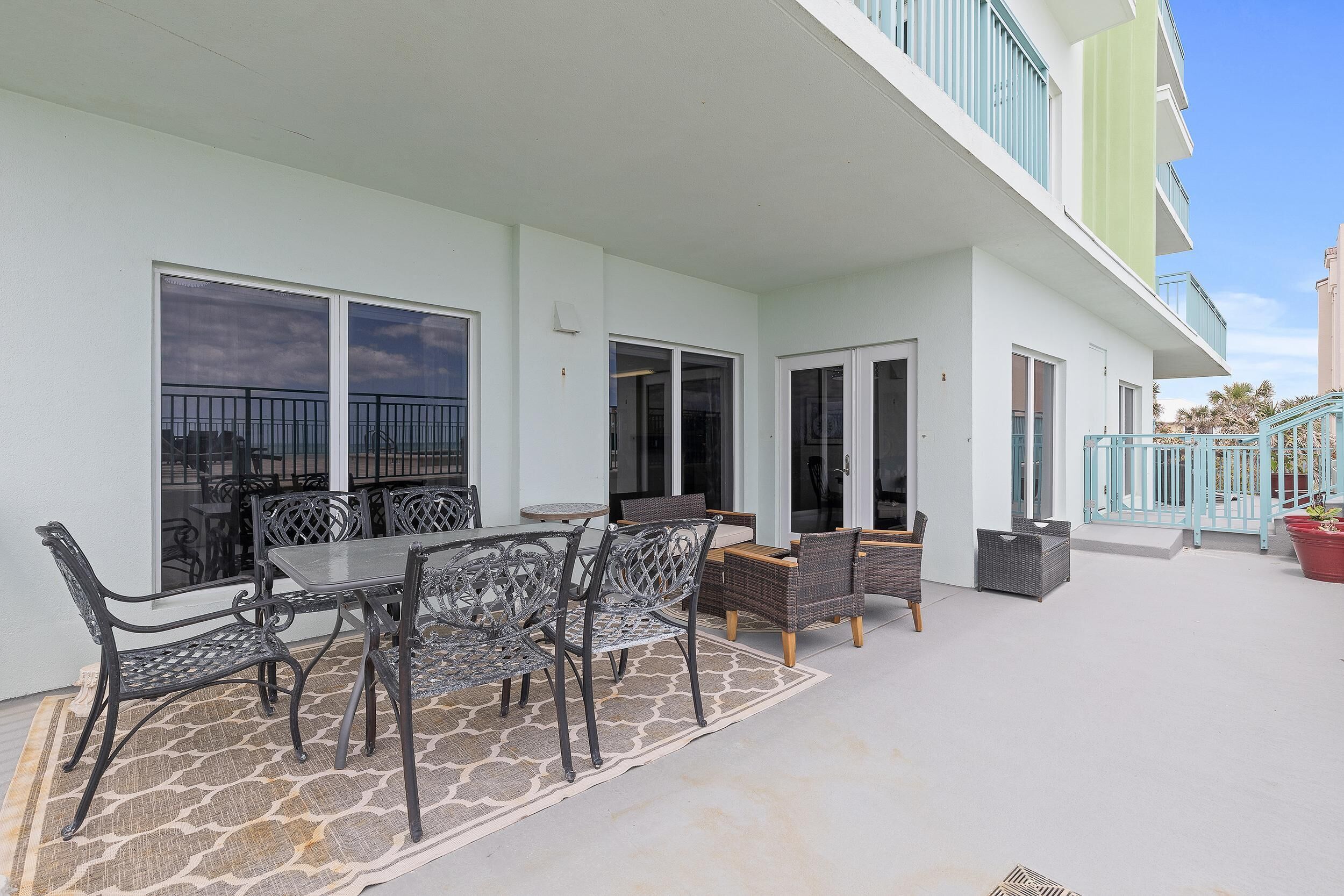 Une terrasse extérieure couverte, aménagée avec un ensemble de mobilier de jardin et des fauteuils en osier, un tapis à motifs de pierre et un bâtiment doté d’un balcon vert clair.