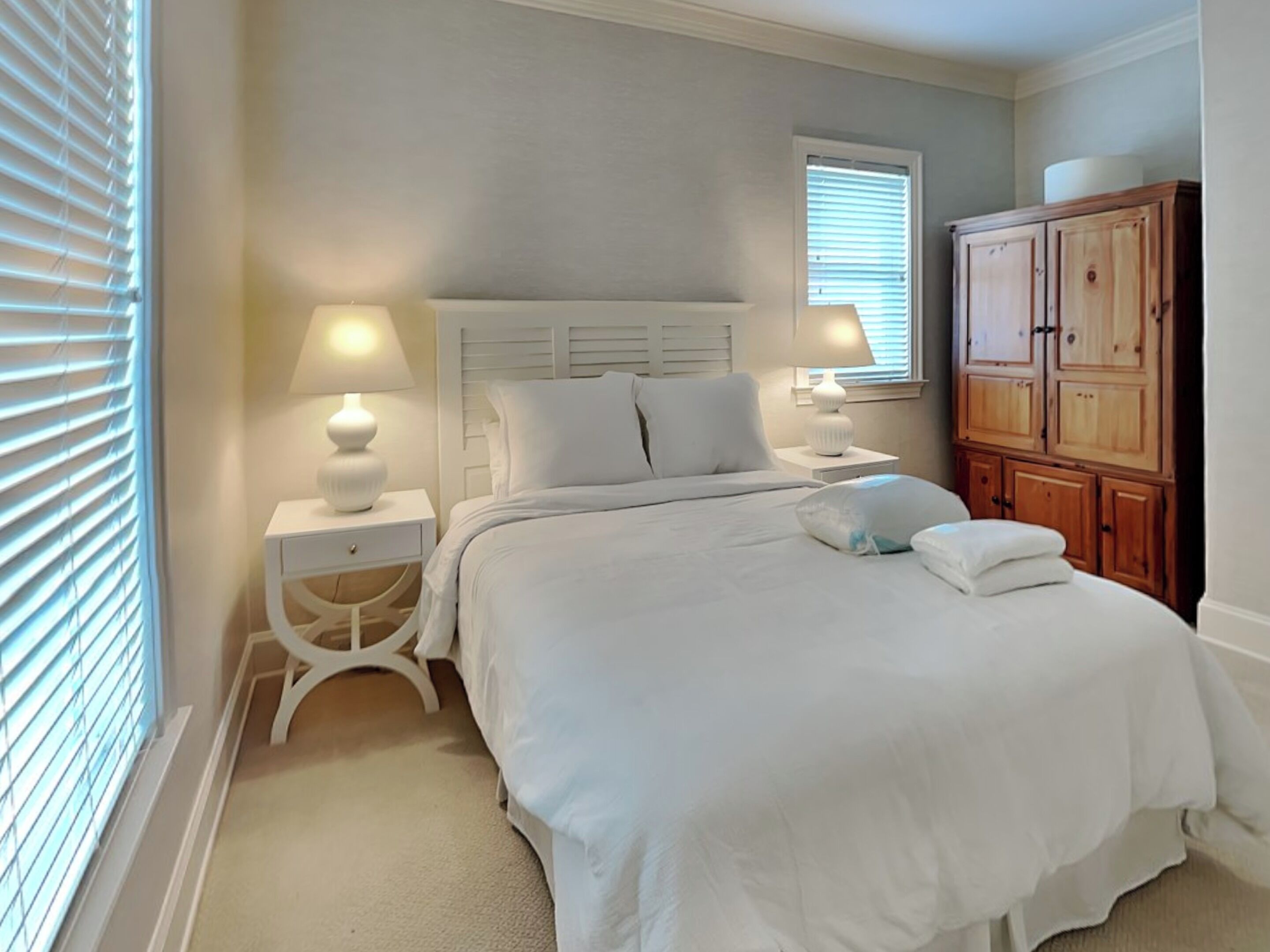 A bedroom with a bed, two bedside tables, a nightstand, a window with blinds, and a wooden wardrobe.