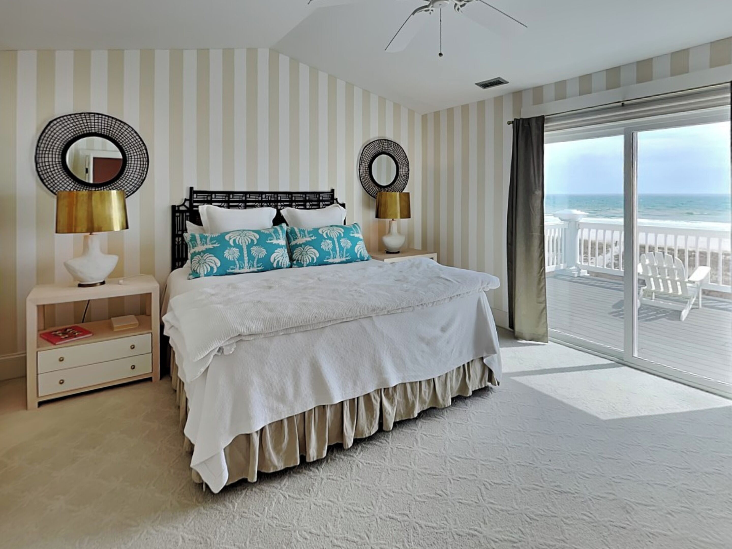 A bedroom with a bed, two nightstands, two wall-mounted lamps, a large window with a view of the beach, and a ceiling fan.