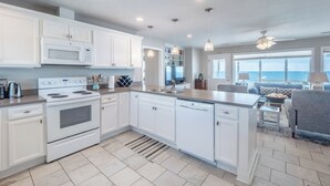 A modern kitchen with white cabinetry, a built-in oven, and a dishwasher. There is a large island with a sink and a dining area with a view of the ocean. - The Shark and the Seahorse by Topsail Realty (Holly Ridge)