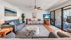 Ein modernes Wohnzimmer mit einem grauen Sofa, einem runden Hocker und einem Deckenventilator. Ein Fernseher, ein kleiner Tisch und ein Sessel sind ebenfalls vorhanden.
