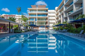 Una piscina con tumbonas y sombrillas, rodeada por un edificio moderno con balcones.