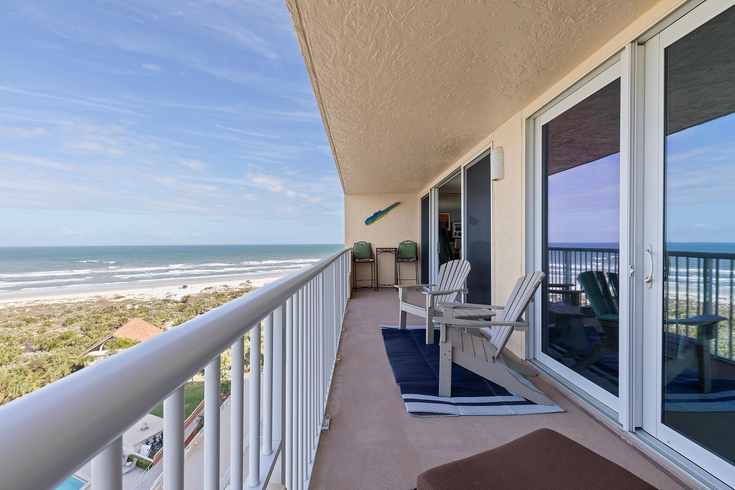 A balcony with a view of the beach, white chairs, and a table.