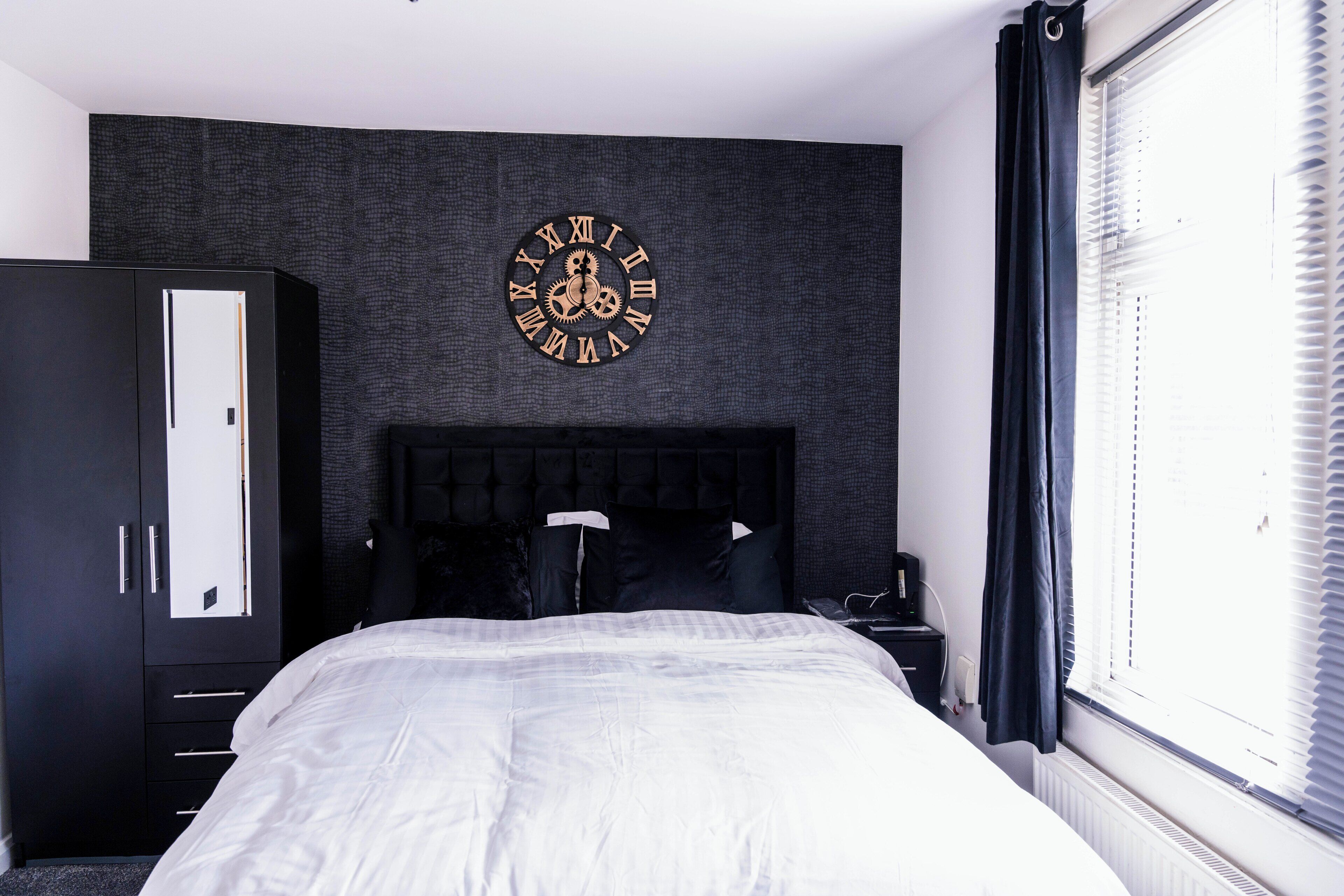 A bedroom with a large bed, a wardrobe, a clock on the wall, and a window with blinds.